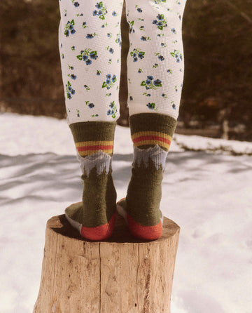 Child wearing floral-patterned leggings and "The Ridge Runner Sock" in Evergreen by THE GREAT. FALL 23 TGO stands on a tree stump with snow-covered ground in the background, ready for an everyday adventure in their luxurious merino wool attire.