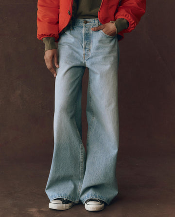 A person wears THE GREAT. F25 D1 The Slouch Wide Leg Jean – Tidal Wash with five-pocket styling, black-and-white sneakers, an olive green top, and a red jacket, standing against a brown background.