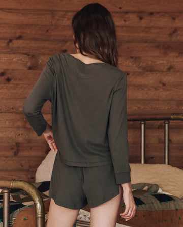 A person with long hair stands with their back to the camera, wearing a Washed Black Crop Sleep Henley from THE GREAT. HOL 24 SLEEP, which has a flattering fit along with shorts, in a room with wooden walls and a metal bed frame.