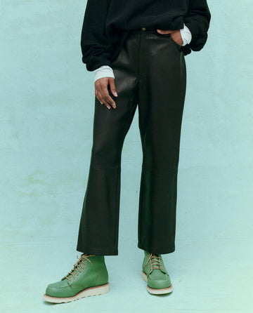 A person wearing The Leather Wayne in black from THE GREAT. HOL 24, with a slouchy fit and hands casually in pockets, stands against a light green background.