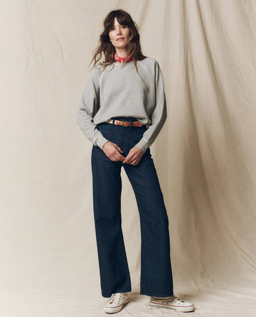 A person stands against a beige backdrop wearing THE GREAT. CORE KNITS's The College Sweatshirt in Varsity Grey, paired with blue wide-leg jeans and white sneakers. They casually tuck their hands into their pockets while a red bandana adorns their neck.