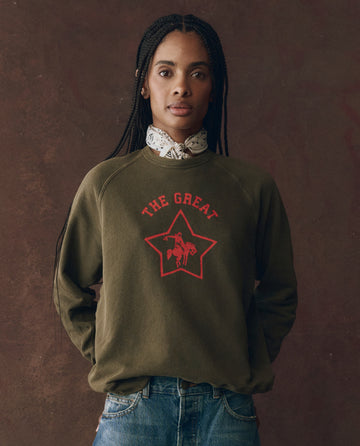 A person wears THE GREAT. F25 D1 The College Sweatshirt in Dark Army with a Rodeo Star graphic, standing against a brown backdrop with hands in pockets.