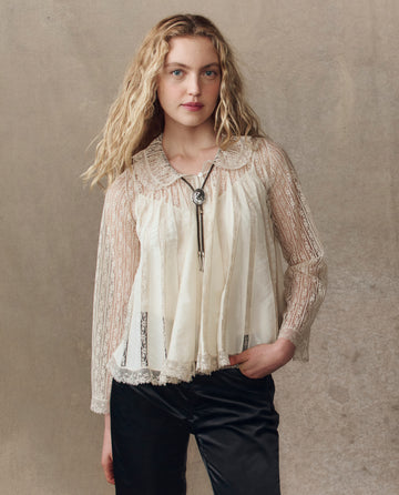 A woman with long wavy blonde hair wears THE GREAT. H25 COLLECTION's The Keepsake Lace Collared Top in cream, styled with a bolo tie and black pants, standing against a neutral textured background.