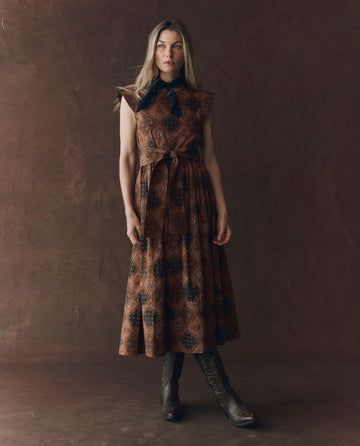 A woman stands against a textured brown background wearing THE GREAT. F25 D1's The Tulsa Dress in Geo Bandana Print, styled with a black necktie and dark cowboy boots.