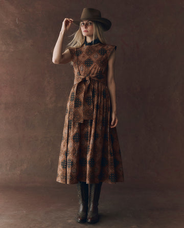 A person wears THE GREAT. F25 D1 The Tulsa Dress in Geo Bandana Print with cowboy boots and a wide-brimmed hat, standing against a plain brown backdrop and holding the hat in one hand.
