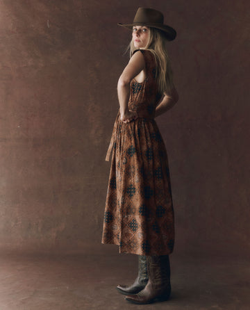 A woman in THE GREAT. F25 D1 The Tulsa Dress—Geo Bandana Print—stands in profile against a plain brown backdrop, wearing a cowboy hat and boots.