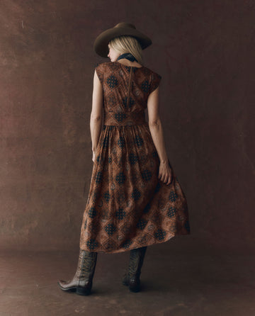 A woman faces away, wearing THE GREAT. F25 D1's The Tulsa Dress in Geo Bandana Print with cowboy boots and a wide-brimmed hat, set against a plain dark background.