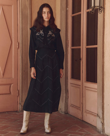 A woman with long dark hair stands in front of wooden doors, exuding fashion-forward elegance in "The Beaded Chevron Skirt" by THE GREAT. SP25 D1, paired with a black top and white boots.