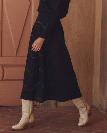 A person in The Beaded Chevron Skirt by THE GREAT. SP25 D1, characterized by its black fabric and white zigzag pattern, paired with beige cowboy boots, walks across a brick surface near a peach-colored door.