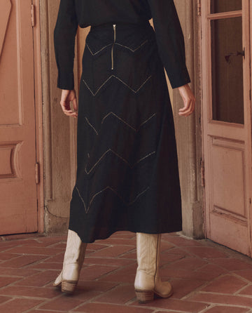 The individual in a black outfit stands facing a wooden door, sporting white cowboy boots and The Beaded Chevron Skirt by THE GREAT. SP25 D1, embodying true fashion.