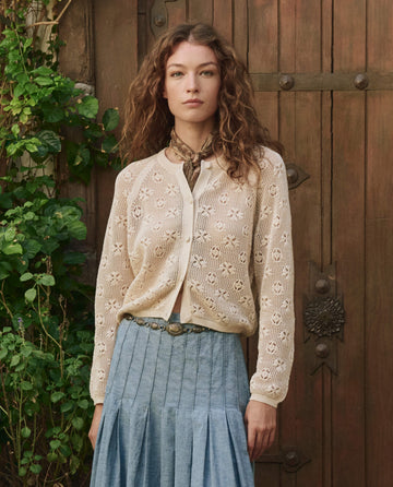 A woman stands by a wooden door, wearing THE GREAT. PF25's The Crochet Cardigan in Natural, styled with a patterned neck scarf and a light blue pleated skirt, with green foliage nearby.