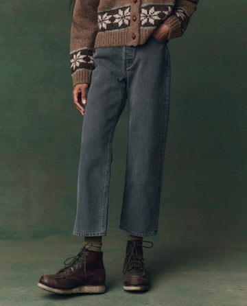 A person wearing a brown patterned sweater, THE GREAT. F25 D2 The Wayne Jean in Graphite Wash with Clean Hem, and dark brown boots stands against a green background.