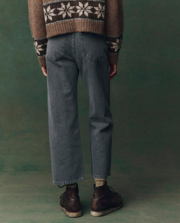 A person stands facing away in THE GREAT. F25 D2’s The Wayne Jean—Graphite Wash with Clean Hem—paired with a brown and white patterned sweater and boots against a green backdrop.