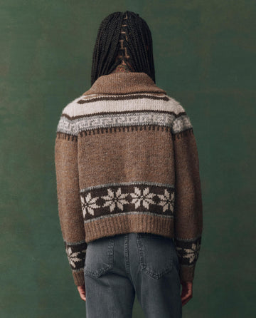 Person with braided hair, shown from behind, wears THE GREAT. F25 D2 The Nordic Scholar Cardigan in Hickory Fair Isle—an oversized sweater with white and black designs—over blue jeans, set against a green background.