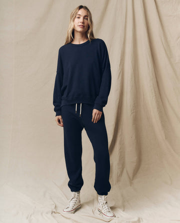 A person stands against a fabric backdrop wearing the True Navy Stadium Sweatpant from THE GREAT. FALL 23 KNITS, a matching sweatshirt, and white high-top sneakers, exuding the charm of vintage athletic wear.