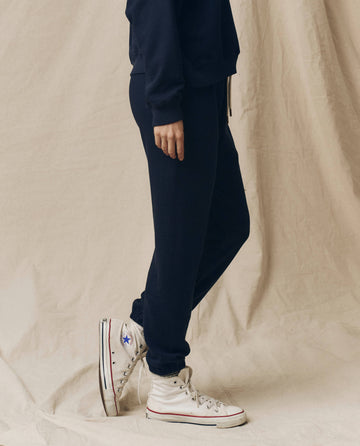 Person wearing The Stadium Sweatpant in True Navy from THE GREAT. FALL 23 KNITS with a matching sweatshirt and white high-top sneakers with dark laces, standing against a light-colored fabric backdrop.