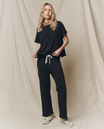 A person stands against a fabric backdrop wearing The Wide Leg Cropped Sweatpant - Almost Black by THE GREAT. CORE KNITS, paired with white sneakers. This loose-fitting outfit features a drawstring waist and is made from 100% cotton.