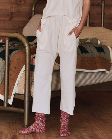 A person wearing The Lounge Crop in Washed White from THE GREAT. HOL 24 SLEEP stands near a bed, hands in pockets. They're accessorized with maroon socks and a cozy set that beautifully complements the patterned quilt.