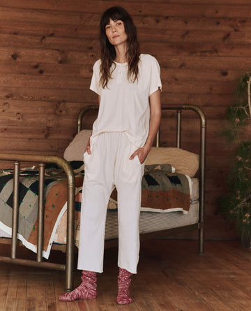 A person stands in a cozy set room with wood-paneled walls, dressed in The Lounge Crop in Washed White by THE GREAT. HOL 24 SLEEP, complemented by pink socks. In the background, a bed adorned with a colorful quilt invites relaxation.
