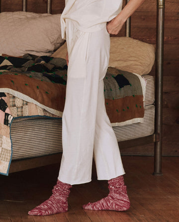 A person dressed in 'The Lounge Crop.' in washed white by THE GREAT. HOL 24 SLEEP stands beside a bed adorned with a colorful quilt and wooden headboard, crafted from super soft jersey, wearing red socks.