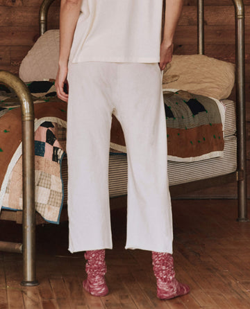 A person stands by a bed draped in a quilted blanket, wearing THE GREAT. HOL 24 SLEEP's The Lounge Crop. in Washed White, complemented by pink socks, forming part of a cozy set in earthy tones within the warm ambiance of a wooden room.