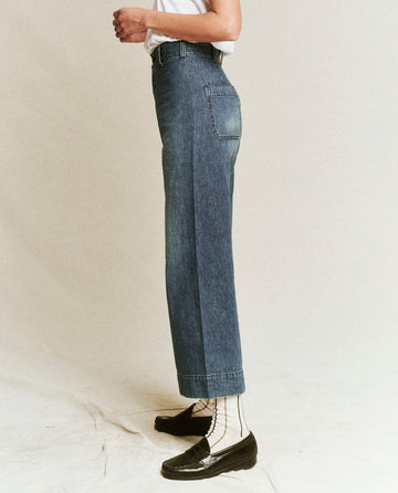 A person stands in side profile, wearing The Seafair Jean - Marina Wash by THE GREAT. PF24, paired with black shoes and a white t-shirt. The background is neutral.