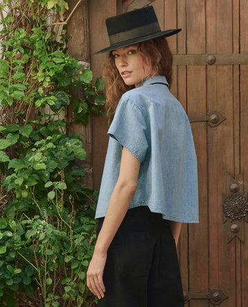 A person in THE GREAT. PF25 The Valor Top -- Boiler Wash, black hat, and black pants stands outdoors near a wooden door and leafy plants, glancing back over their shoulder.