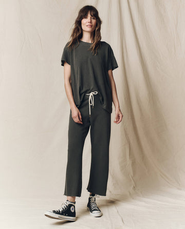 A woman stands against a beige backdrop wearing a dark green T-shirt, The Wide Leg Cropped Sweatpant in Washed Black by THE GREAT. CORE KNITS, and black high-top sneakers.