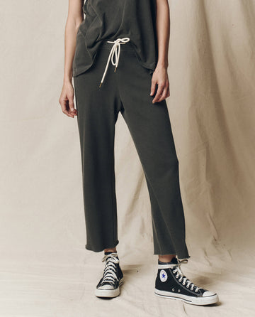 A person in "The Wide Leg Cropped Sweatpant - Washed Black" from THE GREAT. CORE KNITS and black high-top sneakers stands before a beige backdrop.