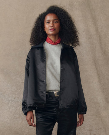 A woman faces forward against a beige background wearing THE GREAT. H25 COLLECTION's The Satin Coach's Jacket in black over a grey shirt, paired with black pants and a red bandana around her neck.