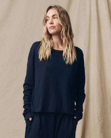 A woman with long blonde hair, wearing The Long Sleeve Crop Tee in True Navy by THE GREAT. FALL 23 KNITS along with dark pants, stands against a beige backdrop, looking to her right.