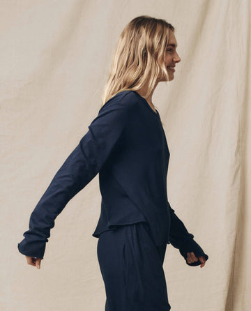 A person with long blonde hair, wearing a handcrafted true navy Long Sleeve Crop Tee from THE GREAT. FALL 23 KNITS, stands against a beige backdrop, facing to the side and slightly smiling.