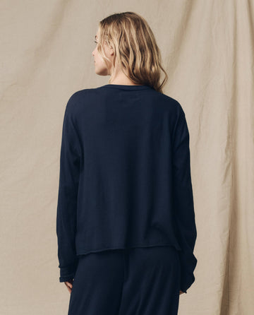 A person with long hair wearing THE GREAT. FALL 23 KNITS' The Long Sleeve Crop Tee in True Navy stands with their back to the camera against a neutral backdrop.