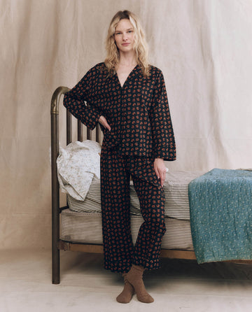 A person stands beside a metal-frame bed, wearing THE GREAT. F25 SLEEP The Pajama Shirt in Navy Horse Medallion Print, brown socks, and surrounded by neutral and teal bedding and blankets against a neutral background.