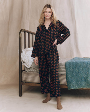A person stands beside a metal-frame bed, wearing THE GREAT. F25 SLEEP The Easy Sleep Pant in Navy Horse Medallion Print, paired with brown socks and bedding in neutral and teal tones. The backdrop is plain beige fabric.