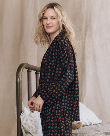 A woman with blonde hair wears THE GREAT. F25 SLEEP’s The Pajama Shirt in Navy Horse Medallion Print, standing by a brass bed with a floral pillow against a neutral backdrop.