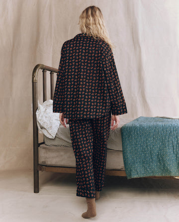 A person with long blonde hair, seen from behind and wearing THE GREAT. F25 SLEEP The Easy Sleep Pant in Navy Horse Medallion Print and brown socks, walks toward a vintage-style bed with patterned 100% cotton bedding.