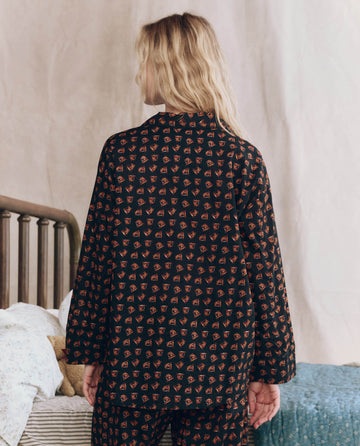 A person with long blonde hair stands facing away, wearing THE GREAT. F25 SLEEP's The Pajama Shirt in Navy Horse Medallion Print, near a bed with patterned bedding.