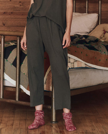 A person wearing The Lounge Crop in Washed Black from THE GREAT. HOL 24 SLEEP stands near a metal bed adorned with a patterned quilt and pillow, with maroon socks adding a pop of color on the wooden floor.