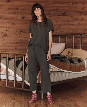 A woman stands in a room with a wooden wall and a metal-framed bed, wearing The Lounge Crop in Washed Black by THE GREAT. HOL 24 SLEEP and red socks. She is beside a quilted bedspread and a stuffed bear, perfectly blending comfort with style.