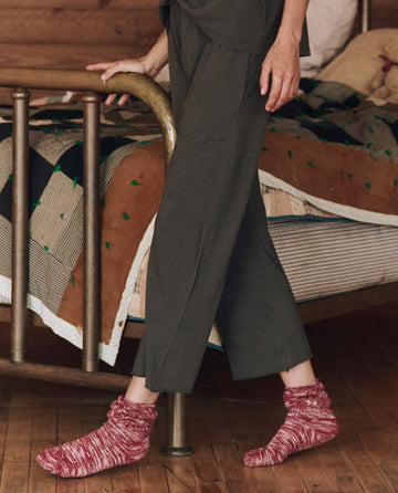 A person in Washed Black Lounge Crop pants stands by a bed adorned with a colorful quilt, where the cozy Sleep Henley from THE GREAT. HOL 24 SLEEP lies folded on the wooden floor beside their red socks.