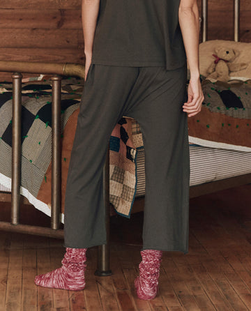 A person stands in a room with a wooden floor, wearing dark loose pants and red socks. In the background, the bed is adorned with a patchwork quilt and cradles a teddy bear. The cozy setting invites relaxation, perfect for lounging in your Sleep Henley or your favorite Lounge Crop by THE GREAT. HOL 24 SLEEP in Washed Black.