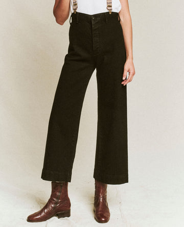A person wearing THE GREAT. F25 D1 The Seafair Jean.. -- Carbon, a white top, suspenders, and brown ankle boots stands against a plain background.