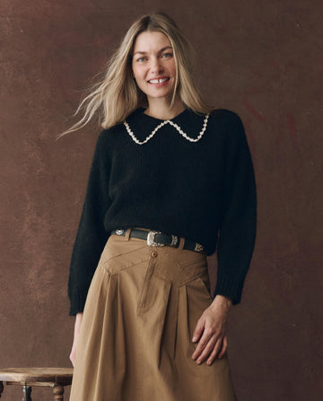 A woman with long blonde hair wears THE GREAT. F25 D1 The Crochet Collar Pullover in black with soft white, styled with a tan pleated skirt, standing before a brown background.