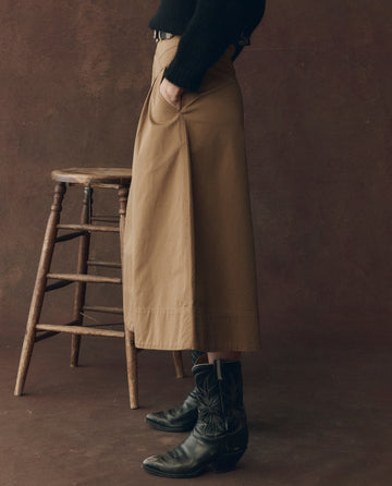A person stands sideways by a wooden stool, wearing THE GREAT. F25 D1 The Field Skirt in Tobacco, a black sweater, and black cowboy boots, set against a brown background.