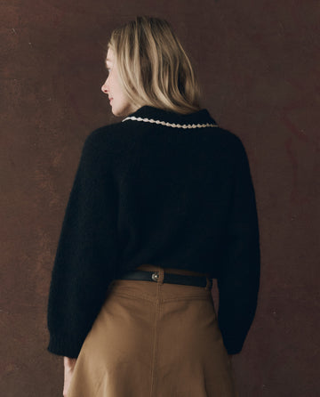 A person with blonde hair, wearing THE GREAT. F25 D1's The Crochet Collar Pullover in Black with Soft White, and a tan skirt, stands facing away from the camera against a dark brown background.