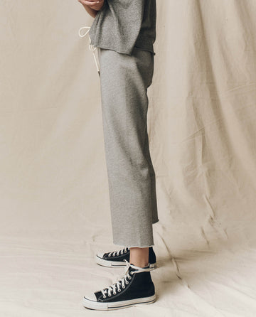 A person stands on a draped fabric backdrop, dressed in The Wide Leg Cropped Sweatpant - Varsity Grey from THE GREAT. CORE KNITS and black high-top sneakers with white soles.