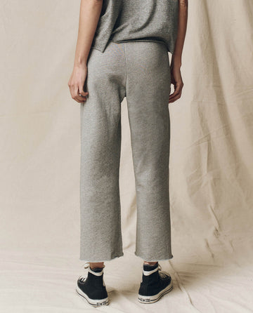 Against a beige fabric backdrop, a person wears THE GREAT. CORE KNITS' "The Wide Leg Cropped Sweatpant" in varsity grey, featuring raw hems and a drawstring waist. The look is completed with stylish black and white sneakers.
