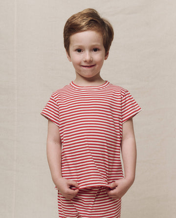 A child wearing The Little Boxy Crew shirt in Sun Cherry Stripe by THE GREAT. SP25 Polo Lounge Little stands against a plain background, smiling slightly while holding the shirt's bottom edge.