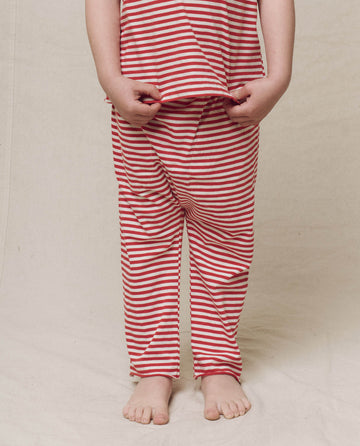A child in The Little Jersey Crop - Sun Cherry Stripe from THE GREAT. SP25 POLO LOUNGE LITTLE stands on a light-colored surface, gently pulling on the shirt like a jersey, with bare feet visible.
